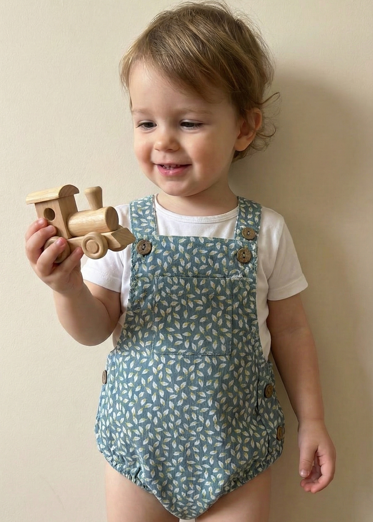 Child wearing a patterned romper holding a wooden toy against a plain background